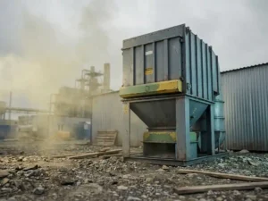 Industrielle Luftfilteranlage auf Baustelle mit gelblichem Dunst aus nahegelegener Verarbeitungsanlage im Hintergrund.