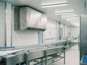 Edelstahl-Luftfilteranlage an weißer Wand einer Lebensmittelproduktionshalle mit Förderband und Fliesenboden darunter.